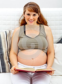 Smiling pregnant woman sitting on sofa with book