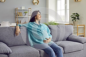 Smiling pregnant woman sitting on sofa at home holding her belly and looking at the window.