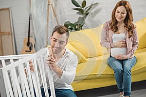 smiling pregnant woman sitting on sofa and looking at happy husband fixing
