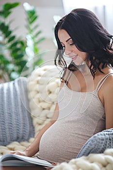 Smiling pregnant woman sitting on sofa and reading magazine