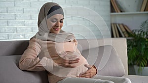 Smiling pregnant woman stroking belly, resting on home sofa, maternity happiness