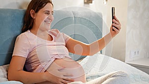 Smiling pregnant woman waiting for baby sitting in bed and calling friend or doctor on video call conference. Pregnant