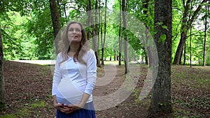 Smiling pregnant woman walks alone in park and strokes her belly.