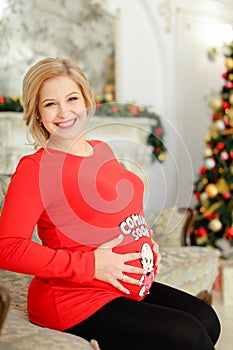 Smiling pregnant woman wearing red sweater hugging bely and sitting on sofa near Christmas tree.