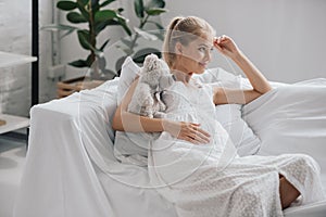 smiling pregnant woman in white nightie with teddy bear resting on sofa