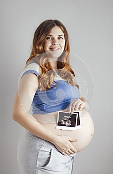 Smiling pregnant young woman with red hair and blue eyes on grey studio background and holding an ultrasound black and white scan