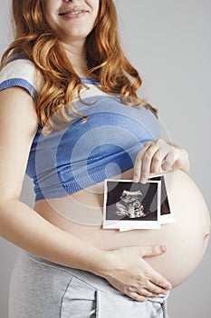 Smiling pregnant young woman with red hair and blue eyes on grey studio background and holding an ultrasound black and white scan