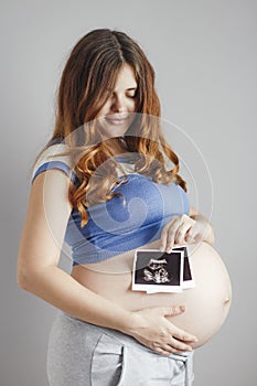 Smiling pregnant young woman with red hair and blue eyes on grey studio background and holding an ultrasound black and white scan