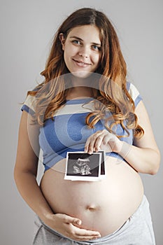 Smiling pregnant young woman with red hair and blue eyes on grey studio background and holding an ultrasound black and white scan