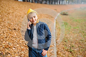 Smiling preteen boy talking on the phone in autumn park. Stylish schoolboy walking after school. People, technology, fashion,
