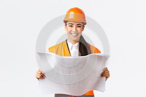 Smiling pretty asian female architect, industrial woman in safety helmet and reflective jacket, being satisfied after