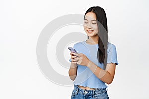 Smiling pretty asian girl using smartphone, holding phone and looking at screen, standing in blue t-shirt over white