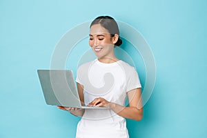 Smiling pretty asian girl in white t-shirt, working on project, looking satisfied and happy at laptop screen