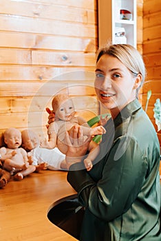 Smiling pretty caucasian young woman holding newborn baby doll in arms while sitting near collection of handmade children toys