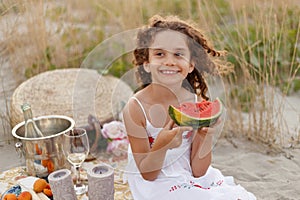 Smiling pretty curly little girl holding a piece of watermelon during summertime vacation at a picnic on the beach