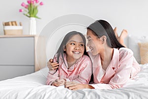 Smiling pretty cute korean millennial woman hugging teenage daughter in pink pajama lie on bed in bedroom