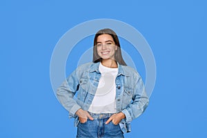 Smiling Latin teen girl wears denim looking at camera stands on blue background.