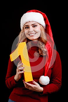 Smiling pretty girl in a christmas hat holding number 1 on a black background. New year 2018 concept.
