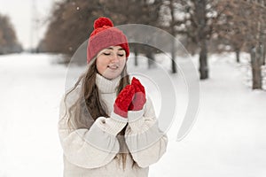 Smiling pretty girl in red knitted hat and mittens and woolen sweater on alley in winter park. Woman outdoor