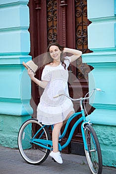 Smiling pretty girl in white dress riding vintage blue bike near beautiful old blue building with antique red doors