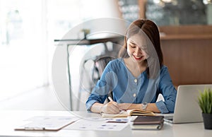 Smiling pretty girl working on laptop computer while sitting at office table, taking notes in a diary.