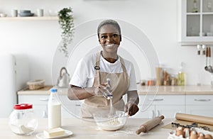 Smiling pretty millennial african american woman chef in apron making dough in light minimalist kitchen interior