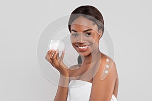 Smiling pretty millennial african american woman with perfect skin in towel with cream on shoulder showing jar