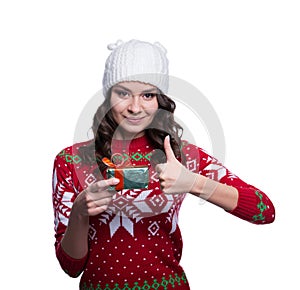 Smiling pretty young woman wearing colorful knitted sweater with christmas ornament and hat, holding christmas gift.