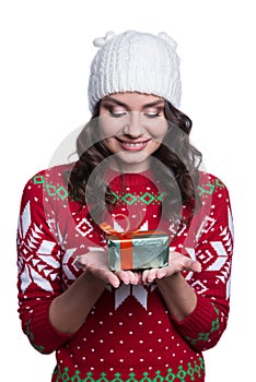 Smiling pretty young woman wearing colorful knitted sweater with christmas ornament and hat, holding christmas gift.