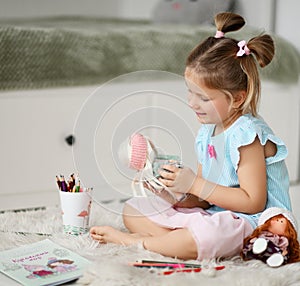 Smiling pretty small girl in home clothing sitting on soft carpet, playing with dolls, drawing with pencils at home