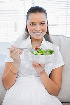 Smiling pretty woman eating healthy salad sitting on sofa