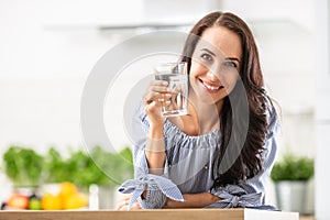 Smiling pretty woman holds a glass of water leaning on kitchen desk