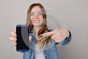 Smiling pretty woman showing black empty screen of her smartphone and pointing on it with her finger; isolated on grey background photo