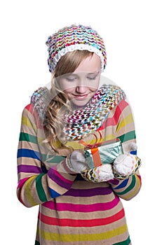Smiling pretty young girl wearing coloful knitted scarf, hat and mittens, holding christmas gift isolated.