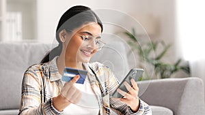 Smiling pretty young Indian brunette girl holding mobile phone and credit card sitting at the table. Happy woman makes