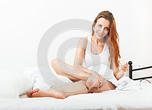Smiling pretty young woman putting cream on legs at home