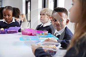 Smiling primary school kids sitting at a table eating their packed lunches together, selective focus