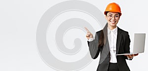 Smiling professional asian female engineer or architect at construction, wearing safety helmet and suit, pointing finger