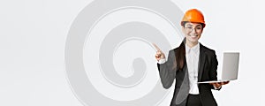 Smiling professional asian female engineer or architect at construction, wearing safety helmet and suit, pointing finger