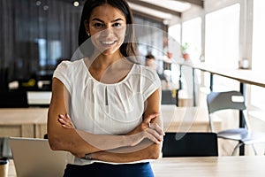 Smiling professional businesswoman in casual, with arms crossed standing in office.