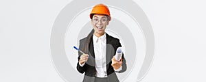 Smiling professional female architect, asian engineer in helmet and business suit, holding blueprints and pen, siging