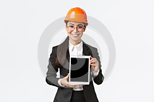 Smiling professional female engineer, saleswoman or estate agent showing location to client, standing in safety helmet