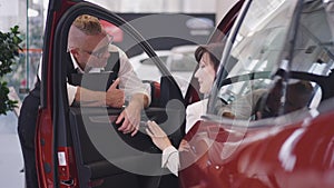 Smiling professional male car dealer with tattoo and mohawk haircut standing at open car with female buyer sitting on