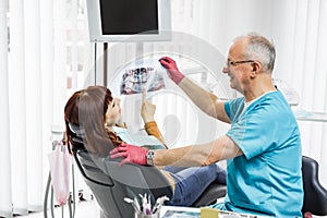 Smiling professional senior male dentist showing dental X-ray to young woman patient, sitting in dental chair in modern