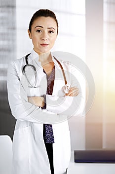 Smiling professional woman-doctor is standing with crossed arms in a sunny clinic office. Portrait of friendly female