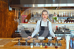 Smiling proud waitress woman standing behind counter looking at camera - Portrait of happy beautiful confident female with apron