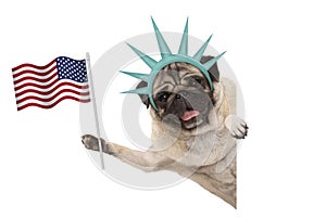 Smiling pug puppy dog holding up American flag, sideways from white banner, wearing lady Liberty crown