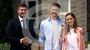 Smiling realtor with documents and happy couple with keys posing near house