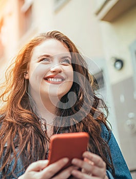 Smiling red curly long hair caucasian teen girl walking on the street and browsing the internet using the modern smartphone.