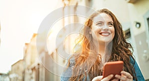 Smiling red curly long hair caucasian teen girl walking on the street and browsing the internet using the modern smartphone.